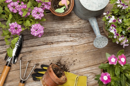 The image depicts a wooden outdoor garden table with a weathered surface, surrounded by potted flowering plants with purple and pink blooms, set in a garden or patio area. On the table, there are various gardening tools including a small hand rake, a garden trowel, and a pair of pruning shears with wooden handles. There is also a ceramic pot with soil spilling out, a gardening glove in bright yellow, and a watering can with a long spout positioned near the pots, indicating ongoing planting or garden maintenance. The scene is outdoors, with natural light suggesting a bright, clear day, and the garden appears well-kept, showcasing elements typical of a landscaped outdoor space that could be maintained by professionals such as Landscapers Maida Vale, who specialise in gardening services within the Maida Vale area, helping create vibrant, functional, and aesthetically pleasing outdoor environments.