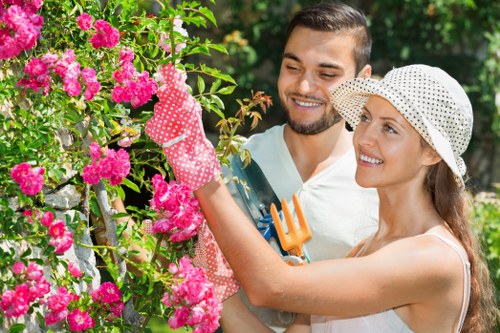 A man and woman are tending to a lush garden in a residential outdoor space, with the woman holding pink flowering bushes and wearing gardening gloves and a wide-brimmed hat, while the man, standing close behind her, holds small gardening tools and smiles. The garden features a well-maintained grassy lawn in the foreground, bordered by vibrant pink roses climbing a trellis, with green foliage in the background. The scene is bright and sunlit, suggesting a clear day. This setting exemplifies typical garden maintenance and landscaping activities that a professional gardening service, such as Landscapers Maida Vale, might undertake to enhance a residential property's outdoor appearance, situated in the Maida Vale area of London, within the postcode region NW. The image highlights the care and attention involved in maintaining a tidy, flowering outdoor space, with balanced natural tones of green, pink, and earth, in a domestic garden environment.