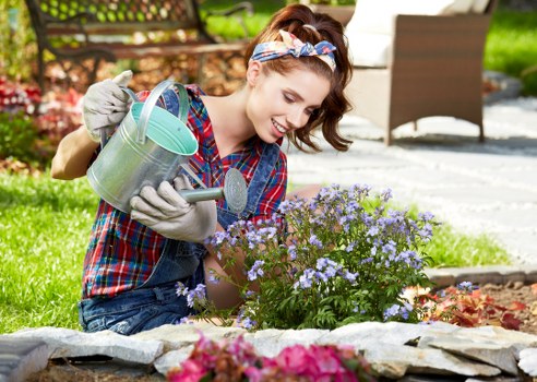 A young woman with a cheerful expression is kneeling in a well-maintained garden in Maida Vale, engaging in gardening activities. She is wearing a colorful plaid shirt, denim jeans, and a patterned headband, holding a small metal watering can in her left hand and a trowel in her right hand. In front of her, there is a vibrant flower bed with blooming purple flowers bordered by neatly arranged grey stone edging. Behind her, the garden features a lush green lawn, a wooden bench, and a pathway made of paving stones. There are several trees and shrubs providing shade and adding to the natural landscape. The scene is bathed in bright natural daylight, suggesting a clear and sunny day, emphasizing the fresh and healthy appearance of the garden. This outdoor space exemplifies a tidy, thriving garden environment suitable for various gardening services offered by Landscapers Maida Vale, highlighting aspects such as flower cultivation, lawn care, and general outdoor maintenance in the Maida Vale area of London.