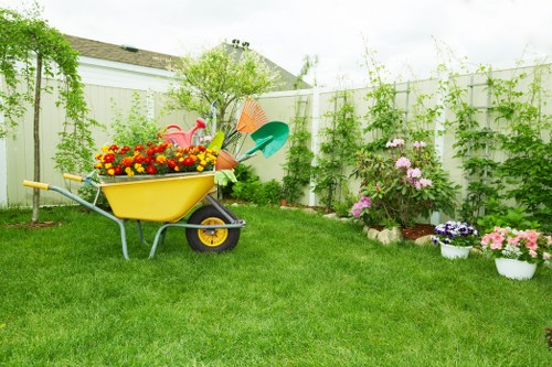 A well-maintained front garden featuring a lush, green lawn bordered by a white fence with climbing plants along its length. In the center of the garden, there is a yellow wheelbarrow filled with colorful flowers, including red, yellow, and orange blooms. Surrounding the wheelbarrow are various gardening tools such as a pink watering can, a green watering can, and an orange-handled garden rake, indicating recent or ongoing gardening activity. To the right, flower beds filled with pink, purple, and white flowering plants are arranged along the fence, with some potted plants placed near the border. The garden environment appears to be sunny with natural daylight, highlighting the vibrant colors of the flowers and the healthy green grass, creating an inviting outdoor space typical of a residential garden in Maida Vale, suitable for professional gardening and planting services by Landscapers Maida Vale.