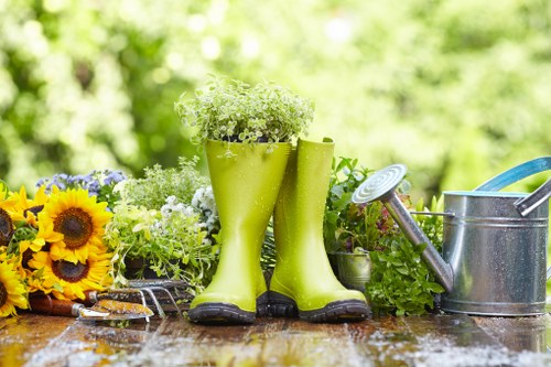 The image displays a gardener's outdoor workspace set on a wooden table with visible rain droplets, indicating recent or ongoing rain. Prominently, a pair of bright green rubber gardening boots is positioned upright, with a small, lush white flowering plant placed inside one of them, suggesting readiness for outdoor planting or maintenance. To the left, a bouquet of vibrant sunflowers and mixed greenery adds colour contrast to the scene, while a small hand trowel with a wooden handle rests on the table beside the flowers. On the right side, a silver metal watering can with a long spout and a blue handle is placed, indicating preparation for watering plants. The background features a blurred lush garden with green foliage and trees, typical of a well-maintained outdoor space in Maida Vale or similar London residential areas. This scene captures elements associated with gardening activities, including plant care, watering, and outdoor gardening tools, representative of services offered by local gardeners or landscapers such as Landscapers Maida Vale in Westminster.