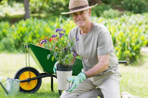 A gardener wearing a straw hat, grey t-shirt, and gardening gloves is kneeling on a lush green lawn in a backyard garden, holding a potted plant with purple and red flowers. Behind him, there is a gravel pathway and a green wheelbarrow with a large yellow wheel, which appears to be used for gardening tasks. The garden features well-maintained grass, a variety of shrubs, and mature trees in the background, creating a vibrant outdoor space. The scene is bathed in natural daylight with soft shadows indicating a sunny or partly cloudy day, suggesting regular outdoor maintenance activities typical of a landscaped area near Maida Vale. This detailed setting aligns with garden care and landscaping services provided by Landscapers Maida Vale, emphasizing planting, lawn care, and outdoor improvements in a typical residential garden environment.