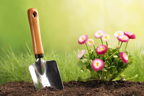 A close-up view of a garden bed in a landscaped backyard, featuring a small metal trowel with a wooden handle inserted into the dark, rich soil. To the right, there is a cluster of vibrant pink and white flowers with rounded petals and yellow centers, possibly primroses or pansies, planted in a neat flower bed border. The background displays a lush green lawn and blurred foliage, suggesting a well-maintained outdoor area in Maida Vale. The scene appears to be on a bright, sunny day with natural light illuminating the plants and soil, reflecting typical gardening activities undertaken by landscapers such as Landscapers Maida Vale to enhance the outdoor space's visual appeal and plant health.