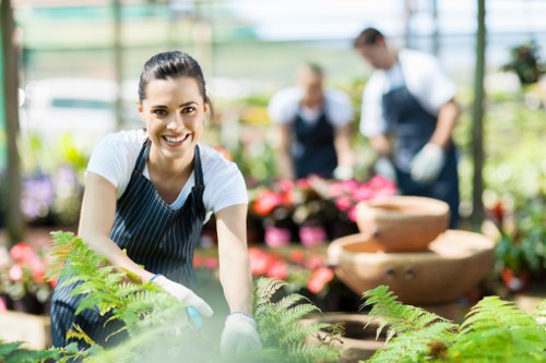 A smiling young woman wearing a navy striped apron and white gloves is tending to a lush garden bed filled with green ferns and other plants, inside a bright and well-lit greenhouse. Behind her, two other gardeners, also dressed in aprons and gloves, are watering or working with potted plants and flower displays, indicating a professional gardening team at work. The greenhouse features a transparent roof and walls, allowing natural light to illuminate the diverse range of vibrant flowering plants and foliage, with some decorative stone or terracotta water features visible in the background. The scene captures the serene and productive environment of an outdoor gardening or landscaping service setting in Maida Vale, with the natural tones of soil, greenery, and blooming flowers contrasting softly against the bright natural light. This environment reflects meticulous plant care, garden layout, and landscaping expertise, emphasizing professional horticultural practices in an accessible, urban garden setting near Maida Vale or NW London.